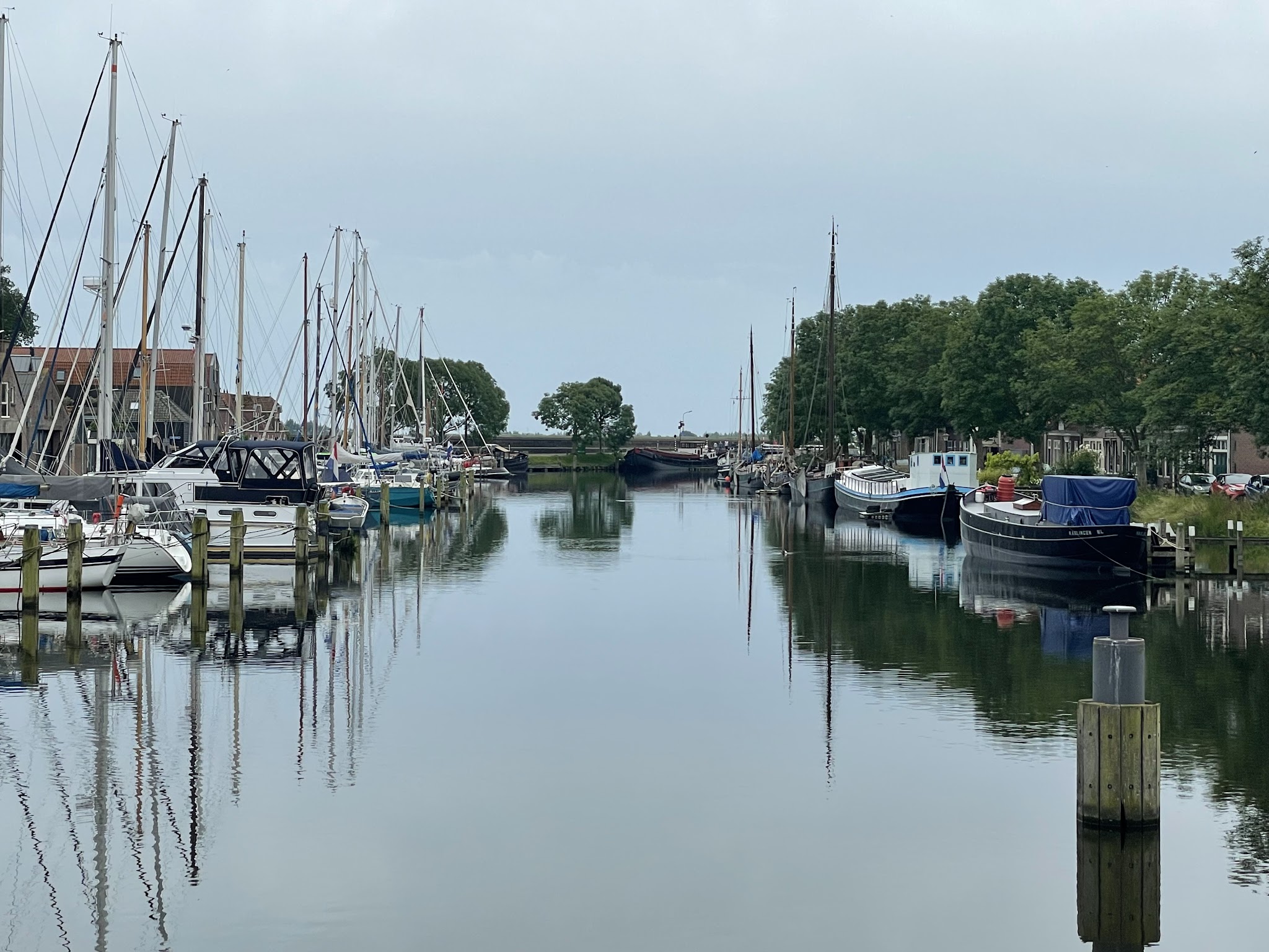 Ruhiger Kanal; Segelboote liegen beidseits an Stegen, Masten spiegeln sich im glatten Wasser unter grauem Himmel.