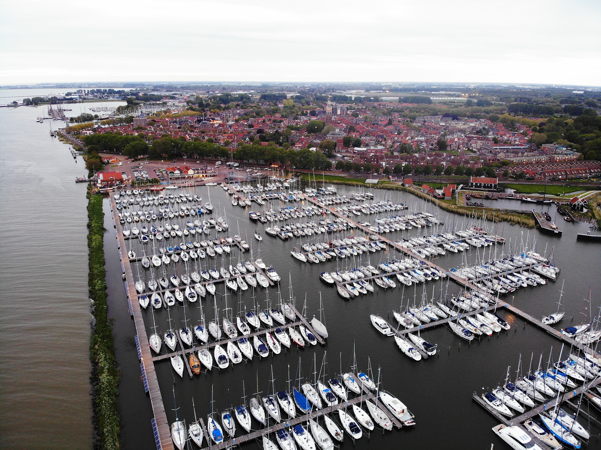 Luftaufnahme eines dicht belegten Yachthafens mit vielen Segelbooten, dahinter eine Küstenstadt am breiten Gewässer.