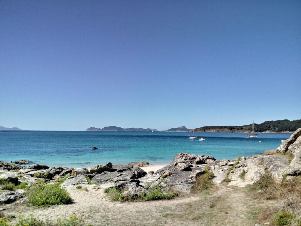 Felsige Küste mit Sandpfad, türkisblauem Meer, drei vor Anker liegenden Segelbooten und Inseln am Horizont.