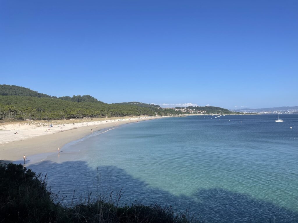 Weite, ruhige Meeresbucht mit hellsandigem Strand, vereinzelten Badenden, bewaldeten Hügeln und klarem Himmel.