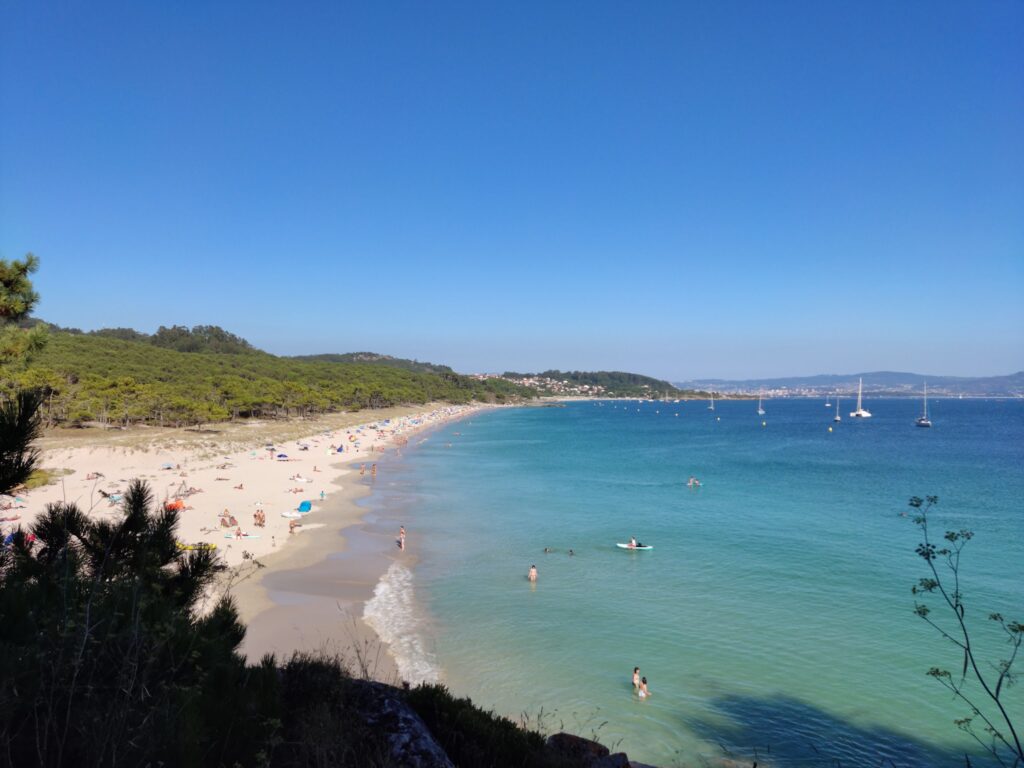 Langer Sandstrand mit Badegästen, türkisblaues Meer, ein paar Segelboote, Pinienwald und klarer Himmel.