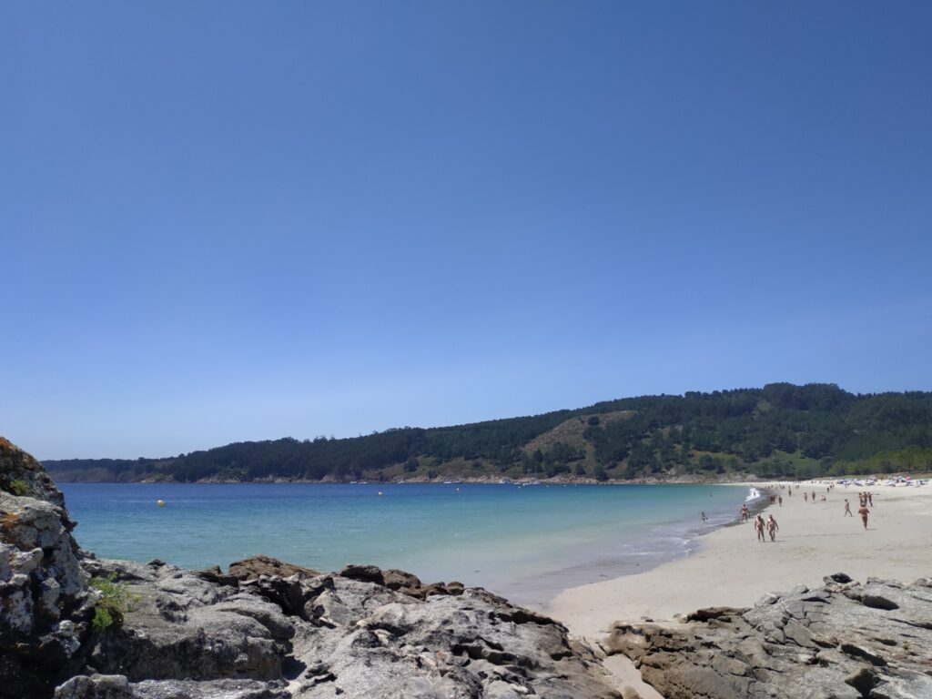 Felsige Bucht mit sandigem Strand, badenden Menschen, türkisblauem Meer und bewaldeten Hügeln unter klarem Himmel.