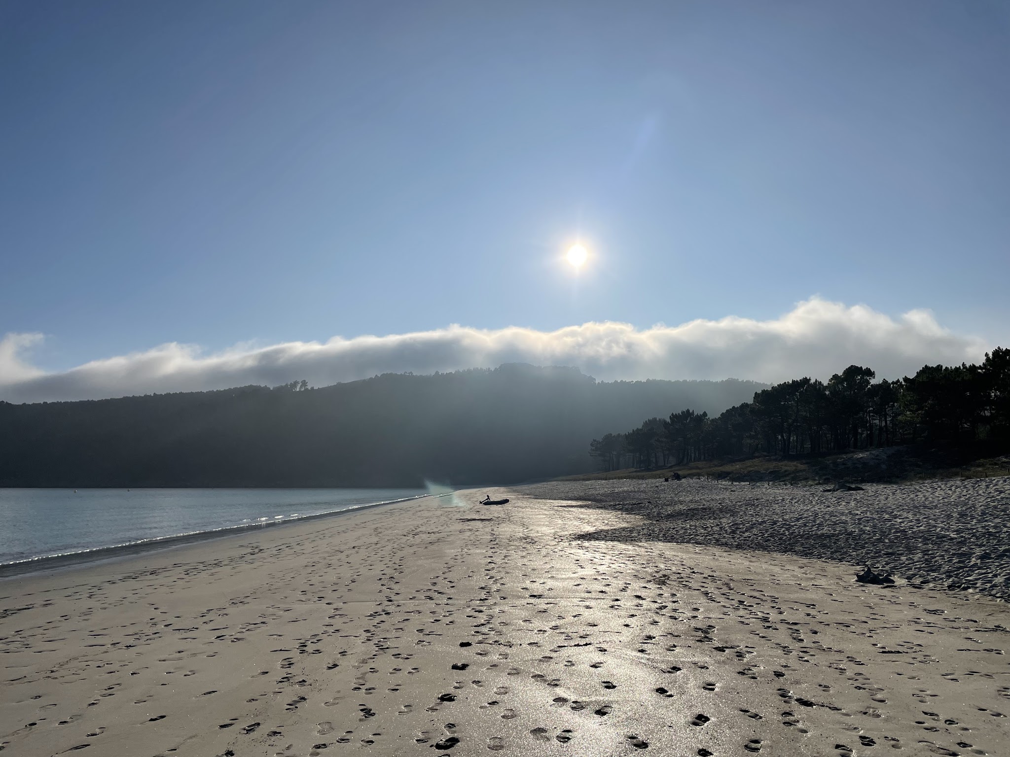 Leerer Sandstrand mit Fußspuren, ruhigem Meer, Wald im Hintergrund, Sonne über tief hängenden Wolken.