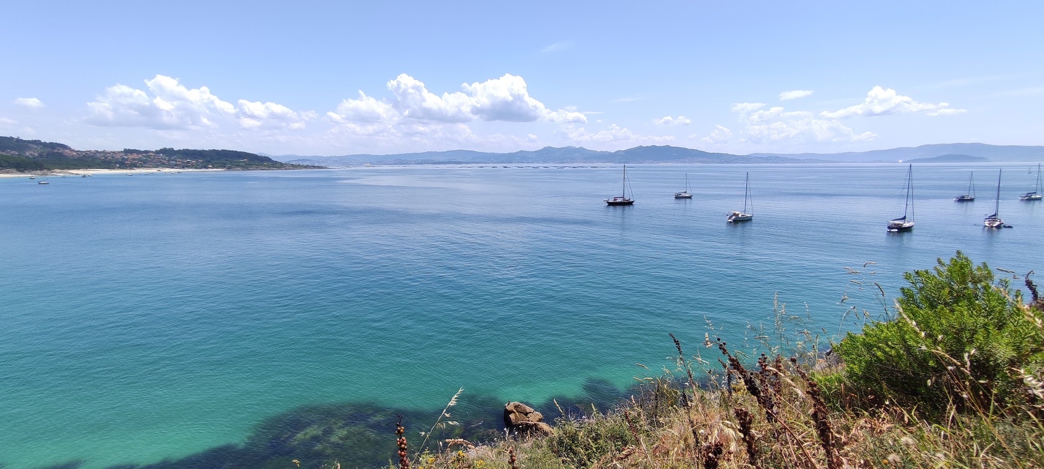 Türkisblaue Bucht, mehrere vor Anker liegende Segelboote, im Vordergrund bewachsene Klippe, Berge am Horizont