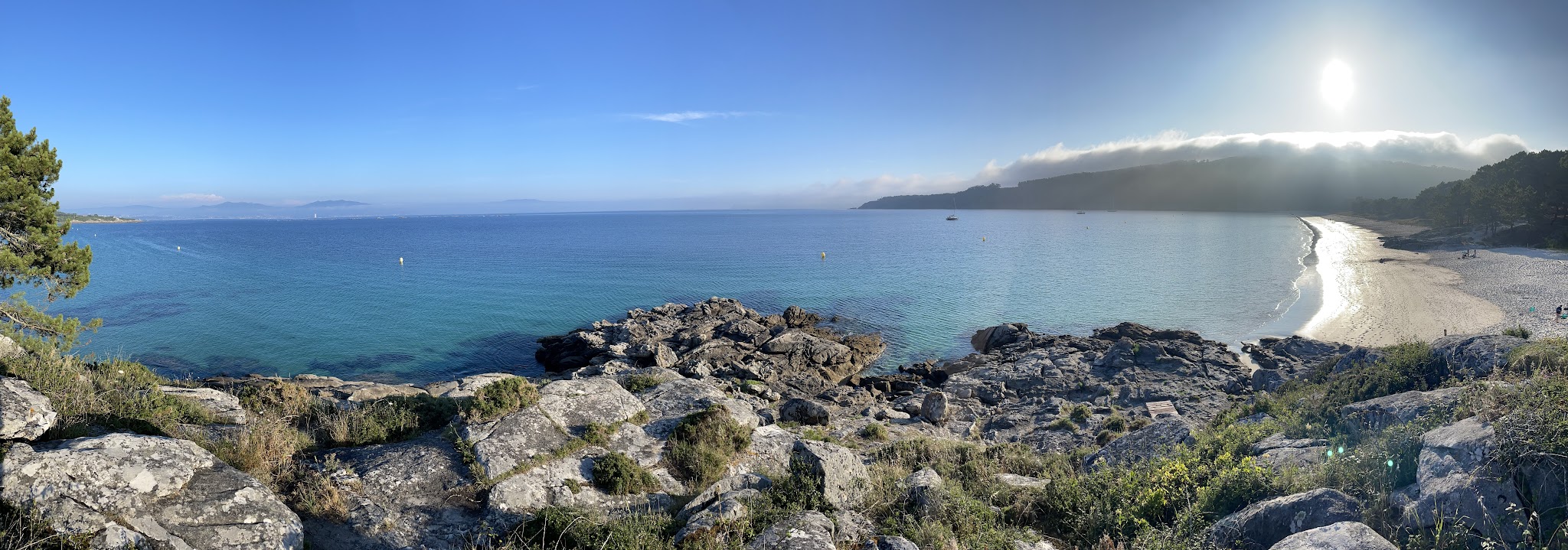 Panorama: türkisblaues Meer, Felsen im Vordergrund, geschwungener Sandstrand rechts, tief stehende Sonne über nebligen Hügeln