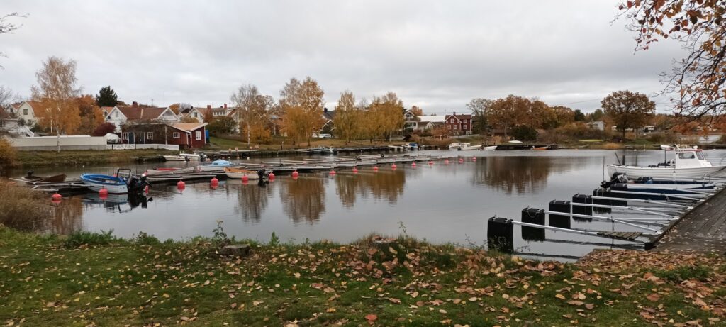 Ruhiger Herbsthafen: kleine Boote an leeren Stegen, bunte Uferhäuser und gelbrote Bäume spiegeln sich im Wasser.