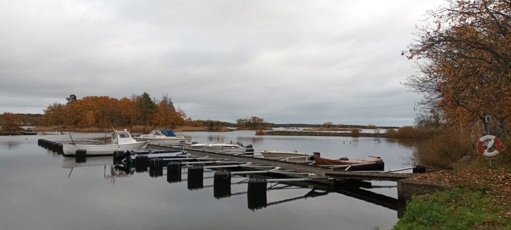 Kleiner Steg mit angelegten Motorbooten in ruhiger Bucht, herbstlich gefärbte Bäume, grauer Himmel.