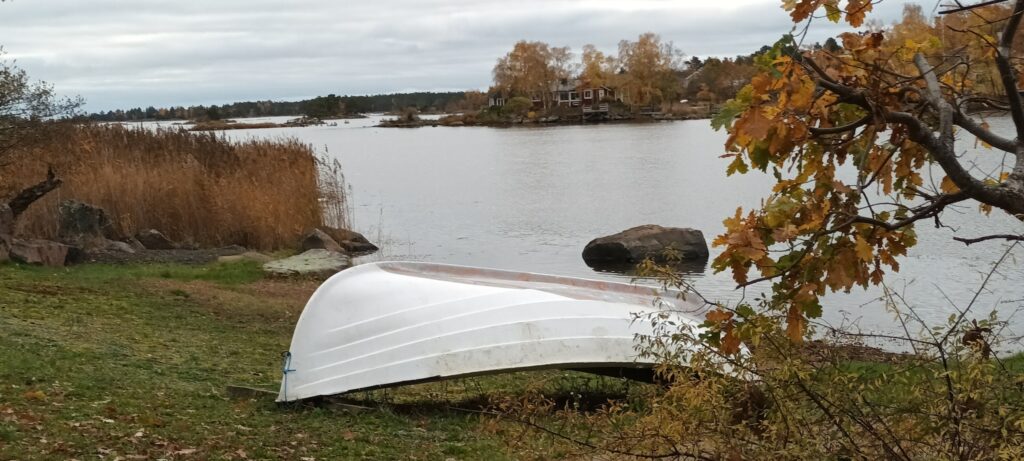 Umgedrehtes weißes Ruderboot am herbstlichen Seeufer, Schilf und ferne Hütte auf kleiner Insel