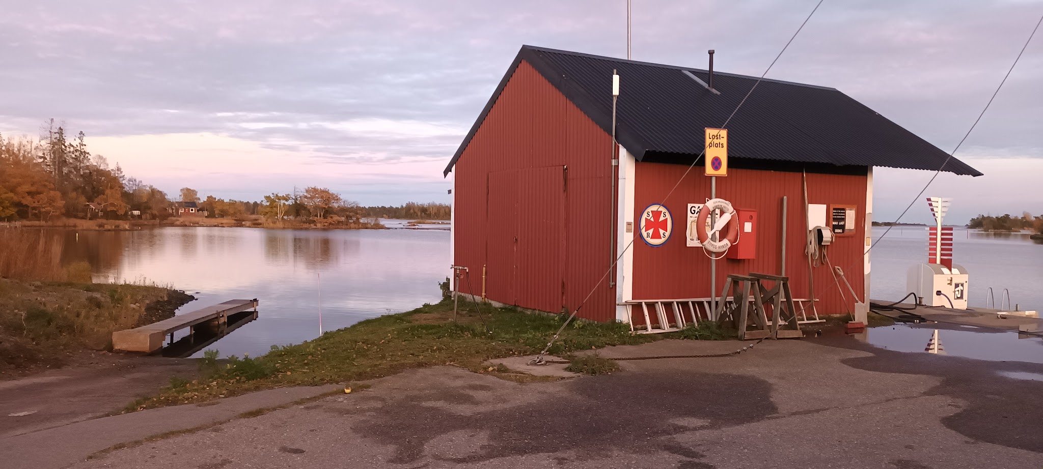 Rotes Bootshaus mit Rettungsring am stillen See; kleiner Steg und herbstliche Bäume im warmen Abendlicht