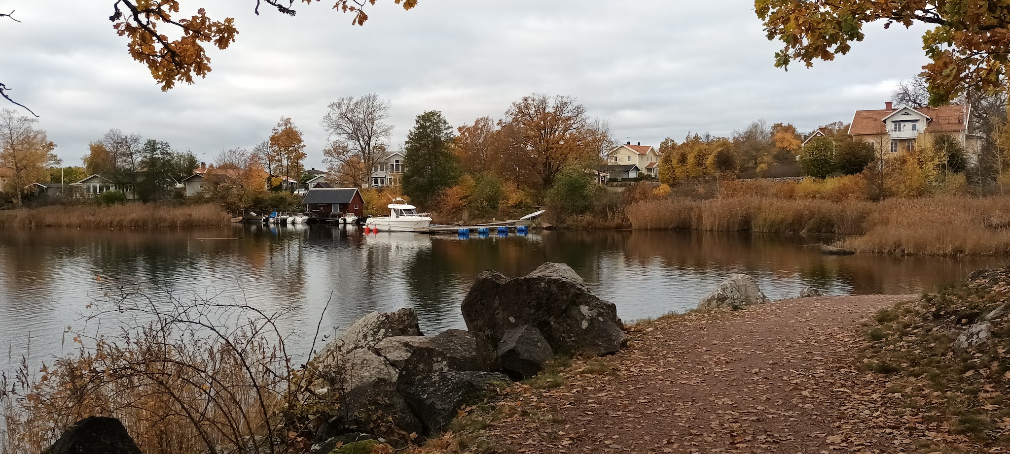 Herbstlicher See mit Laubpfad, Felsbrocken und ruhigem Wasser; Bootssteg mit weißen Booten vor bunten Uferhäusern.