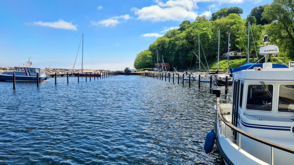 Kleiner Hafen; rechts Motorboot, links Segelboote, mittig Dalben, dahinter bewaldeter Hang unter blauem Himmel