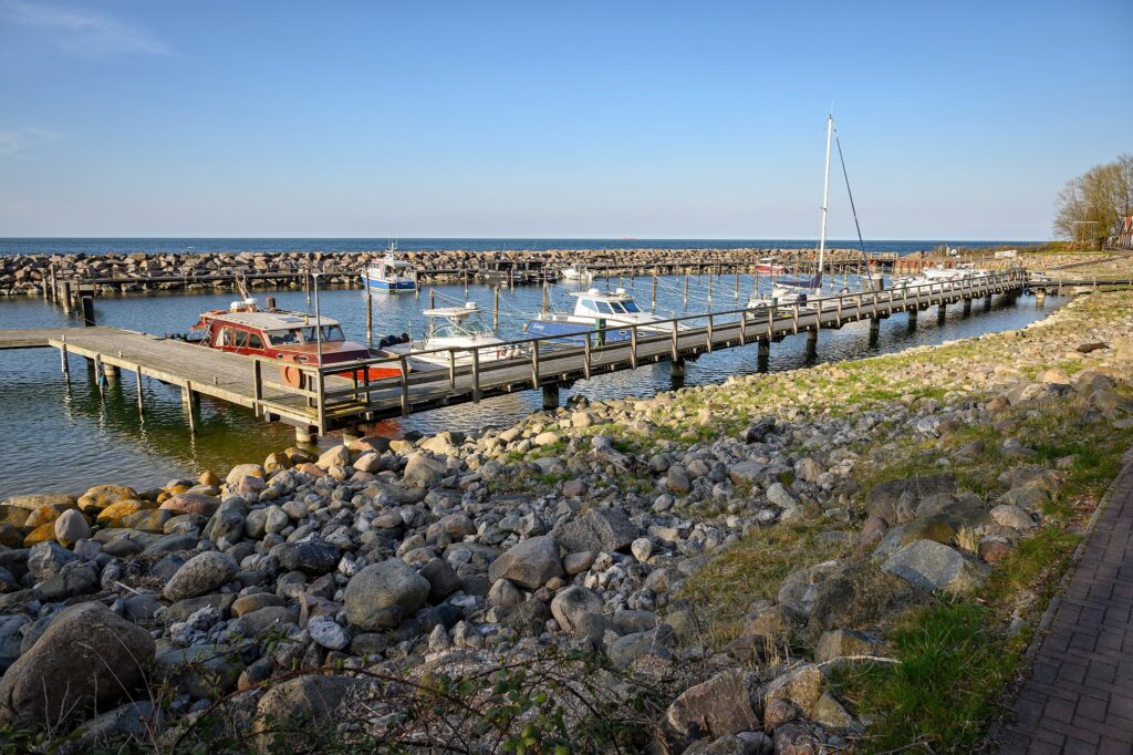 Holzpier mit kleinen Motor- und Segelbooten in ruhigem Hafen, umgeben von felsiger Küste unter klarem Himmel.