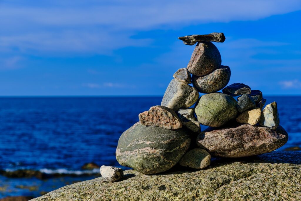 Zu einer Pyramide geschichtete Kiesel balancieren auf Felsen vor tiefblauem Meer und wolkenlosem Himmel.