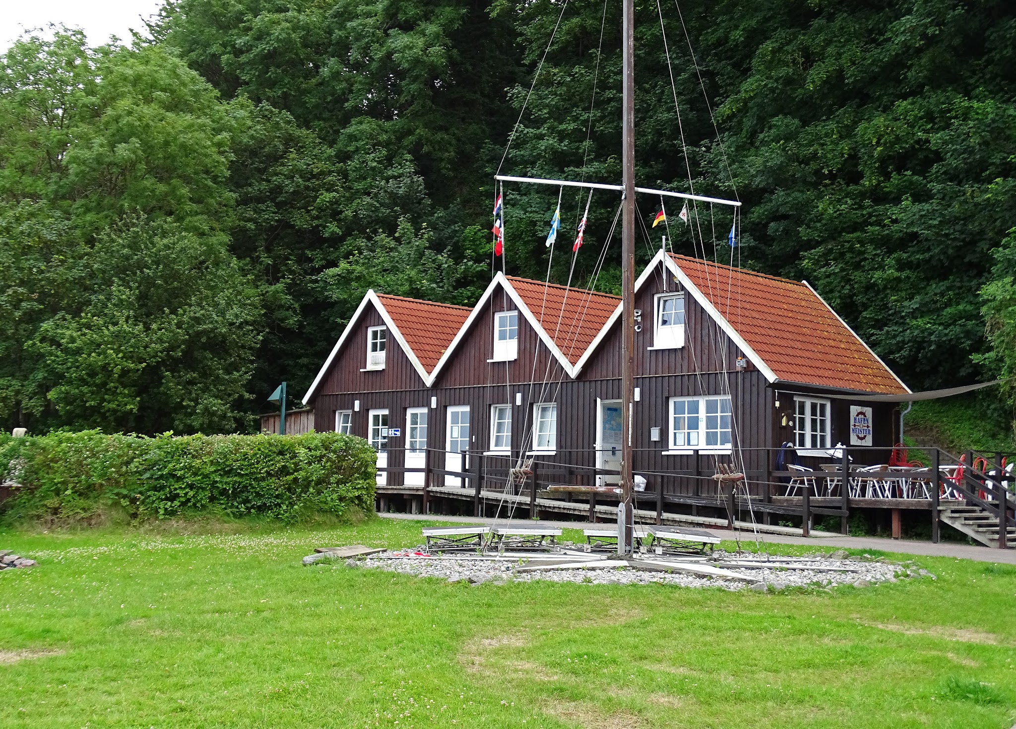 Dunkelbraune Holzhäuser mit roten Giebeldächern, Flaggenmast davor, umgeben von Rasen und dichtem Wald.
