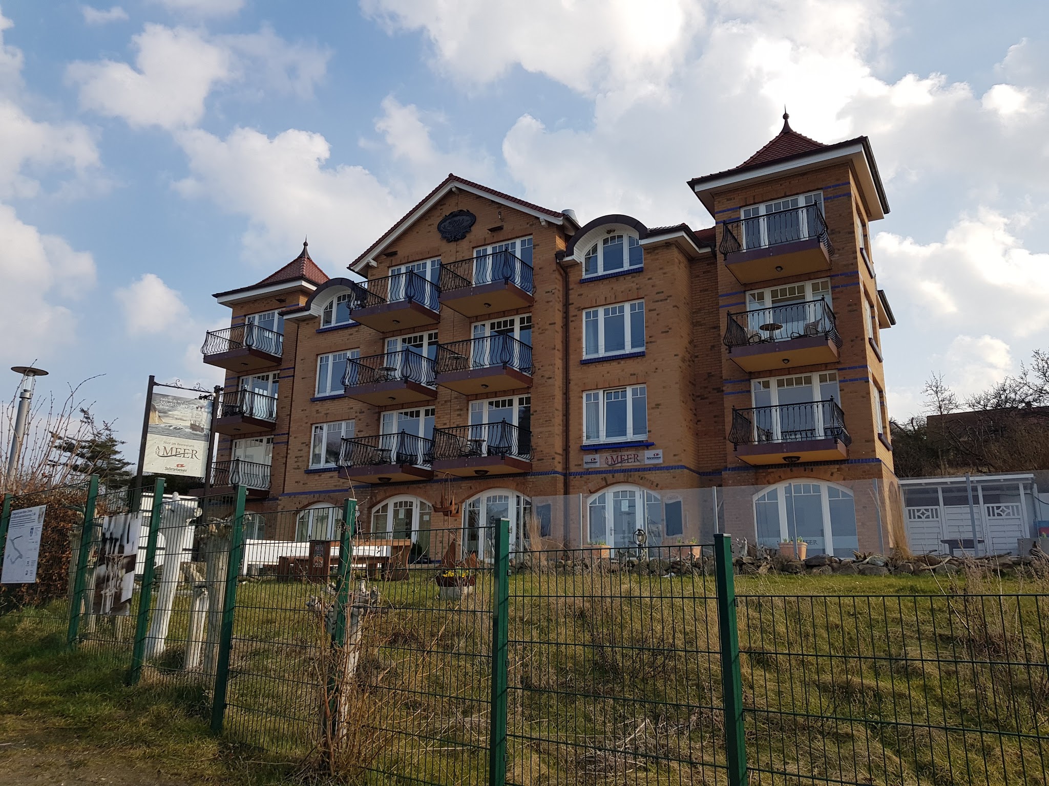 Vierstöckiges Backsteinhaus mit Balkonen und spitzen Türmchen, von Zaun und Rasen umgeben, unter wolkigem Himmel.