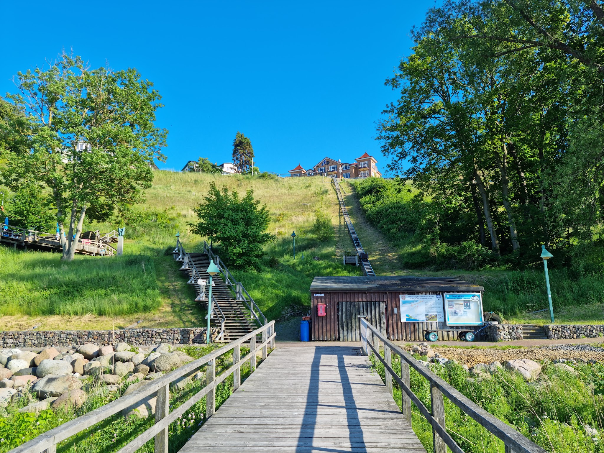 Holzsteg führt zu kleinem Bootshaus; lange Treppe steigt durch grünen Hang zu Häusern auf der Hügelkuppe unter blauem Himmel.