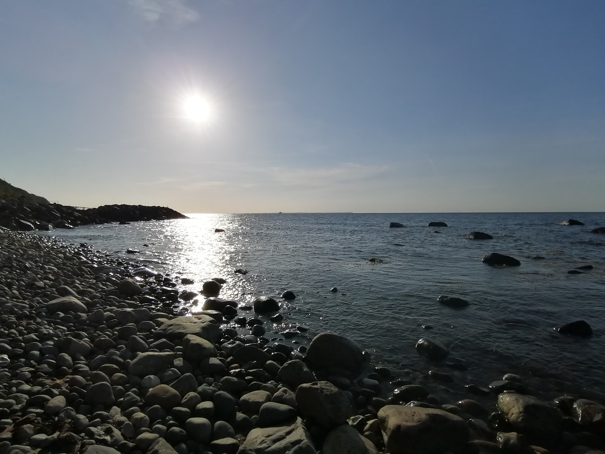 Kiesstrand bei tief stehender Sonne; Licht glitzert auf ruhiger See, vereinzelt ragen Felsen aus dem Wasser.