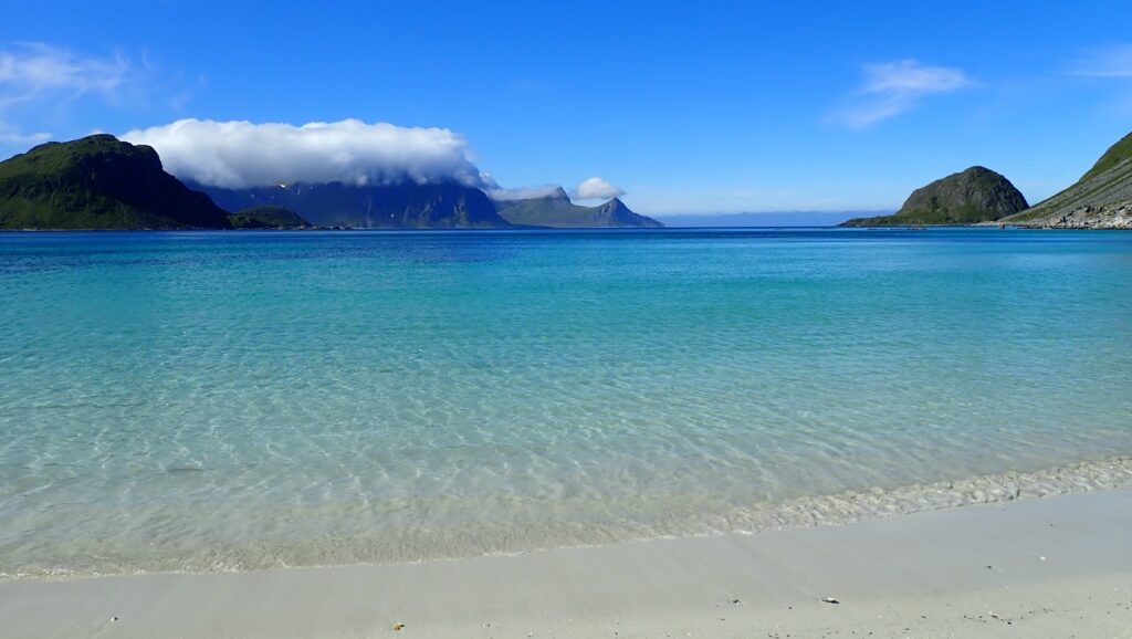 Türkises, klares Meer an weißem Sandstrand; im Hintergrund grüne Berge mit tiefhängender Wolkenhaube unter blauem Himmel