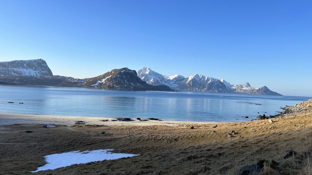 Ruhige Bucht: türkisblaues Wasser, sandiger Strand, schneebedeckte Berge dahinter, wolkenloser Himmel.