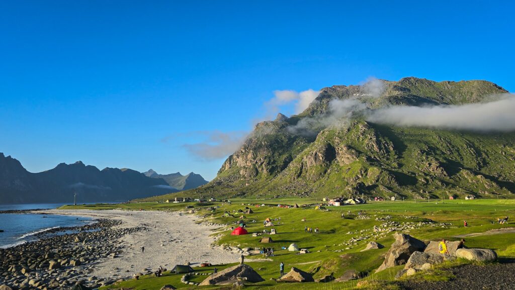 Grüne Bergkulisse an norwegischer Küste, weißer Kiesstrand, verstreute Zelte und Camper bei klarblauem Himmel