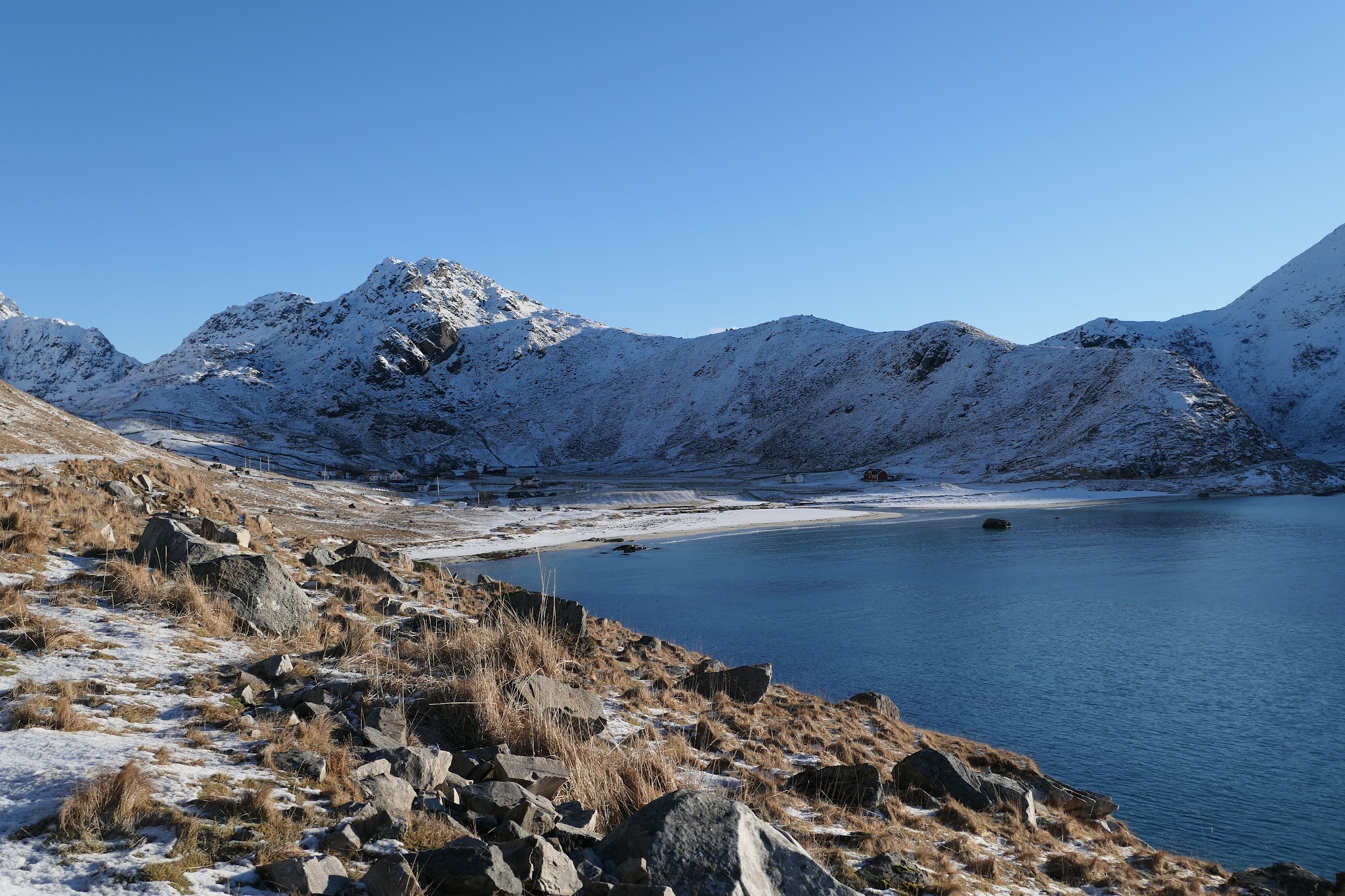 Schneebedeckte Berge hinter ruhigem, tiefblauem See; felsiger, mit trockenem Gras durchsetzter Uferhang im Vordergrund.