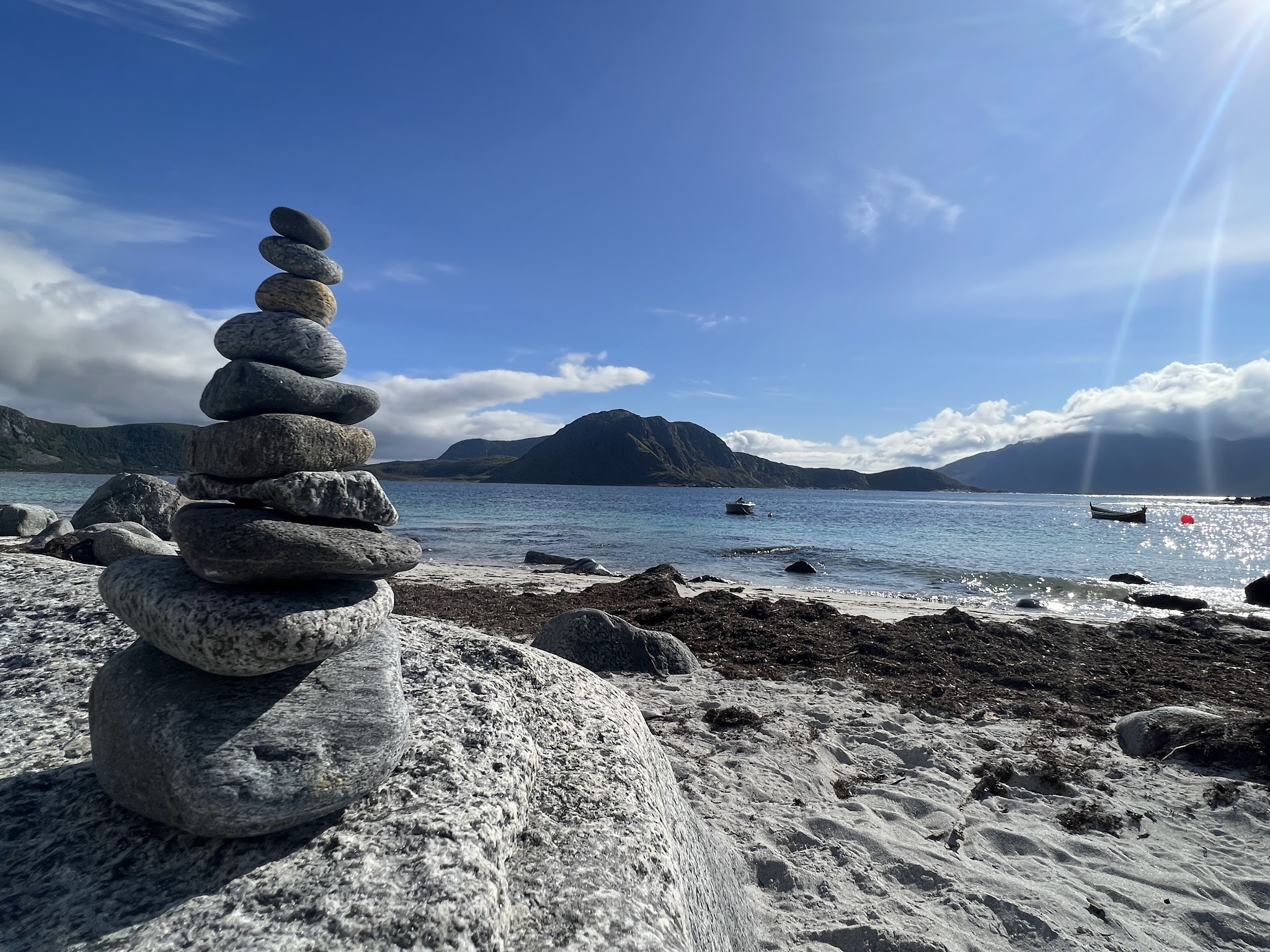 Gestapelte Steine auf felsigem Strand, davor türkisblaues Meer, Boote und sonnenbeschienene Bergkulisse.