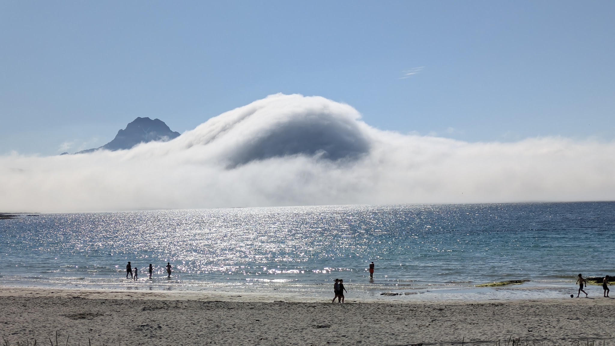 Sonnenbeschienener Strand mit badenden Menschen; glitzerndes Meer, halbverhüllter Berg von kuppelförmiger Nebelwolke umhüllt