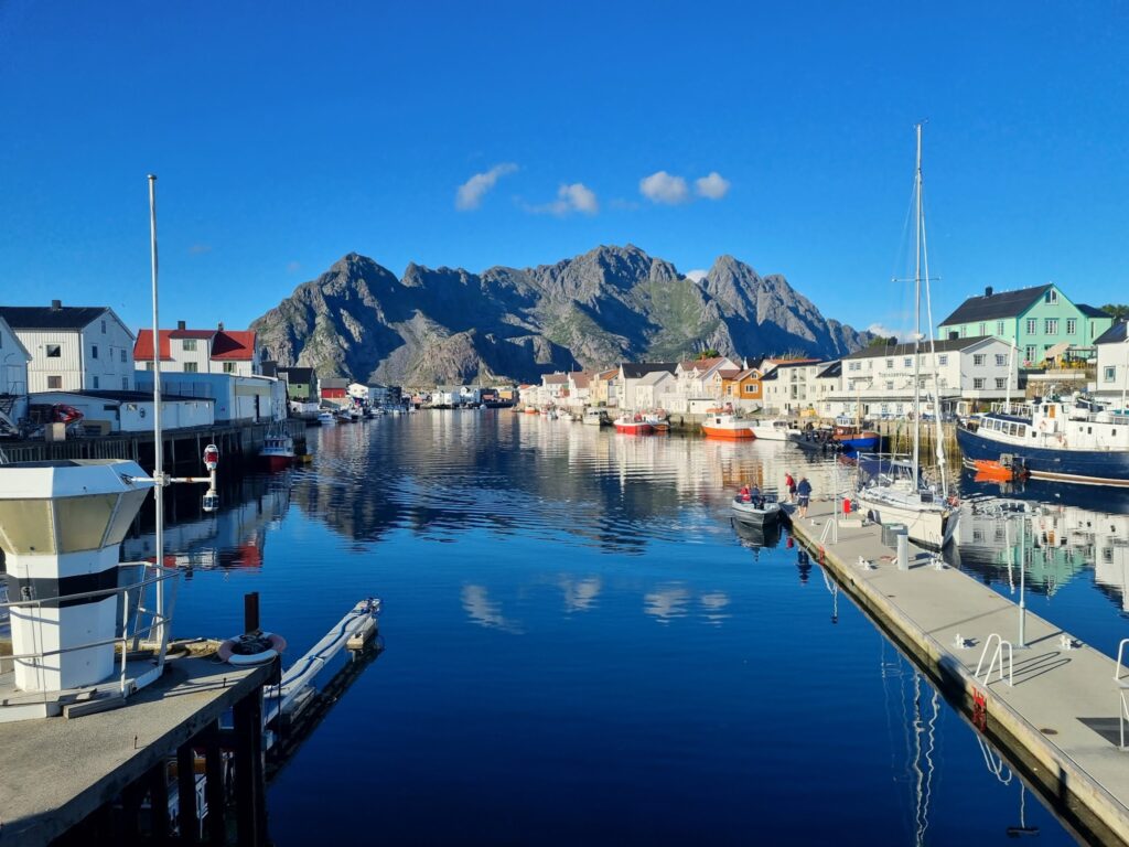 Küstenhafen mit bunten Holzhäusern, Booten und steilen Bergen, die sich im ruhigen, klaren Wasser spiegeln.