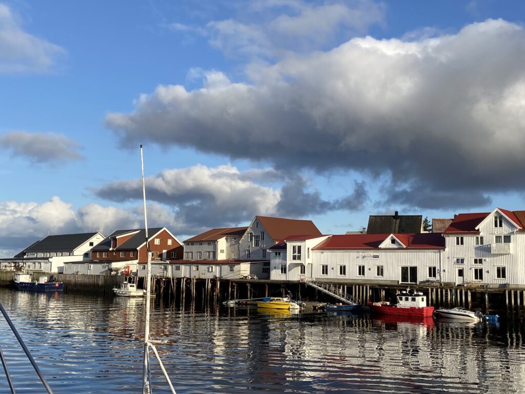 Kleine Hafenhäuser mit roten Dächern spiegeln sich im ruhigen Wasser; bunte Boote liegen am Kai unter Wolkenhimmel.