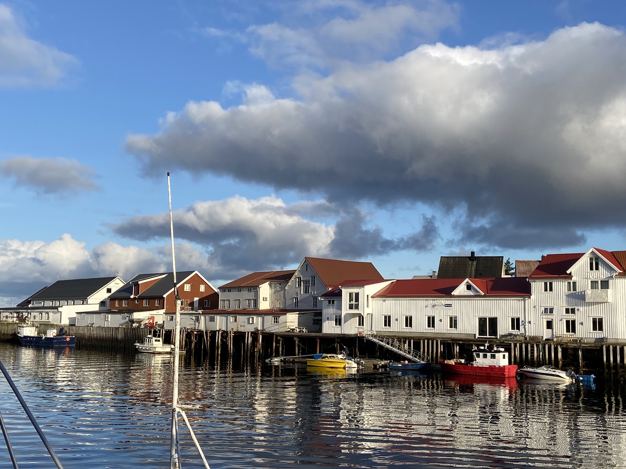 Kleine Hafenhäuser mit roten Dächern spiegeln sich im ruhigen Wasser; bunte Boote liegen am Kai unter Wolkenhimmel.