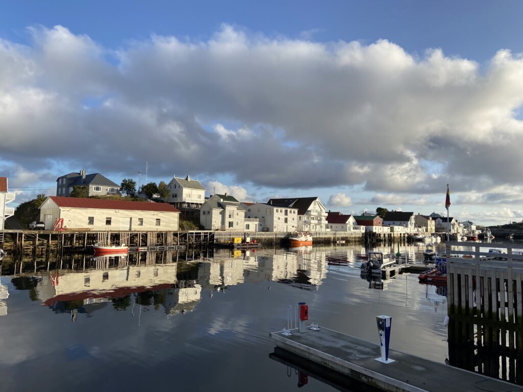 Ruhiger Hafen: weiße Holzhäuser auf Stelzen spiegeln sich im Wasser, kleine Boote vertäut unter wolkigem Himmel.
