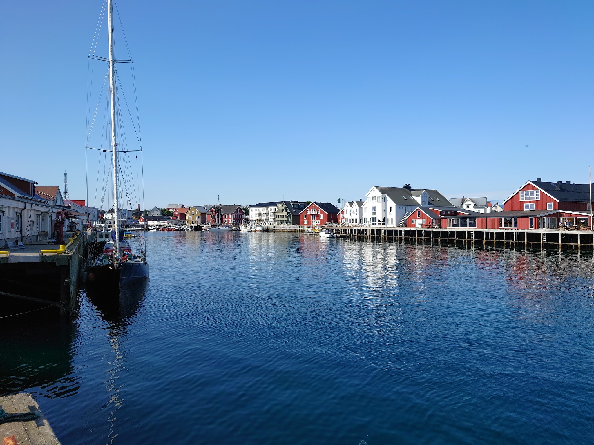 Ruhiger Hafen; links vertäutes Segelboot, rechts bunte Holzhäuser auf Pfählen spiegeln sich im klaren Wasser.