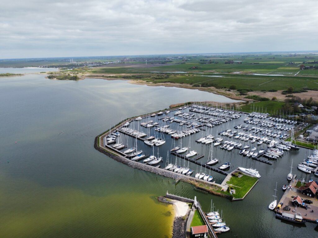 Luftaufnahme eines Yachthafens mit dicht gereihten Segelbooten, von einem Deich und weiten grünen Feldern umgeben.