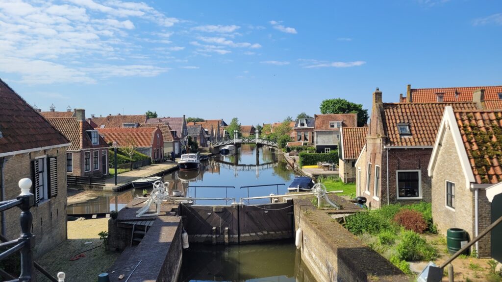 Schleuse vor Backsteinhäusern an ruhigem Kanal mit Booten und Holzbrücke unter blauem Himmel