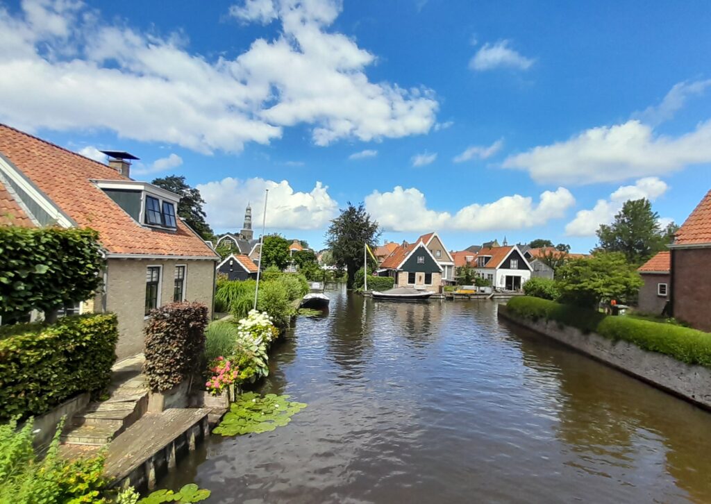 Ruhiger Dorfkanal mit Seerosen, Backstein- und Holzhäusern mit roten Dächern, Boot am Steg, blauer Himmel