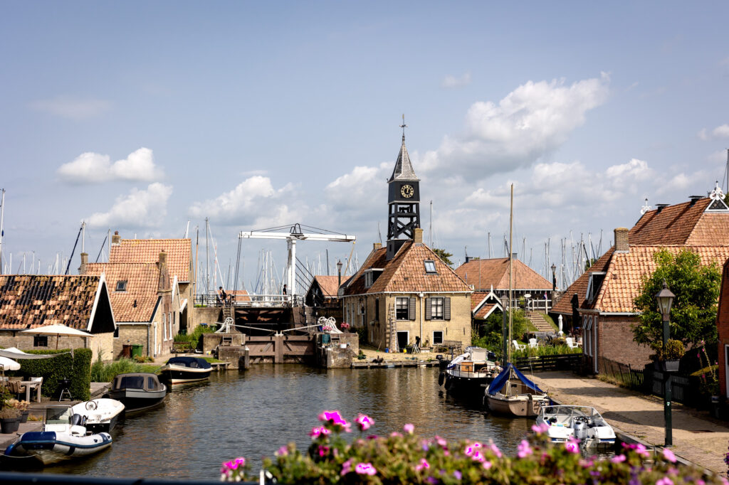 Kleiner Hafenkanal mit Booten, ziegelgedeckte Häuser, Uhrturm und weiße Zugbrücke unter blauem Himmel.