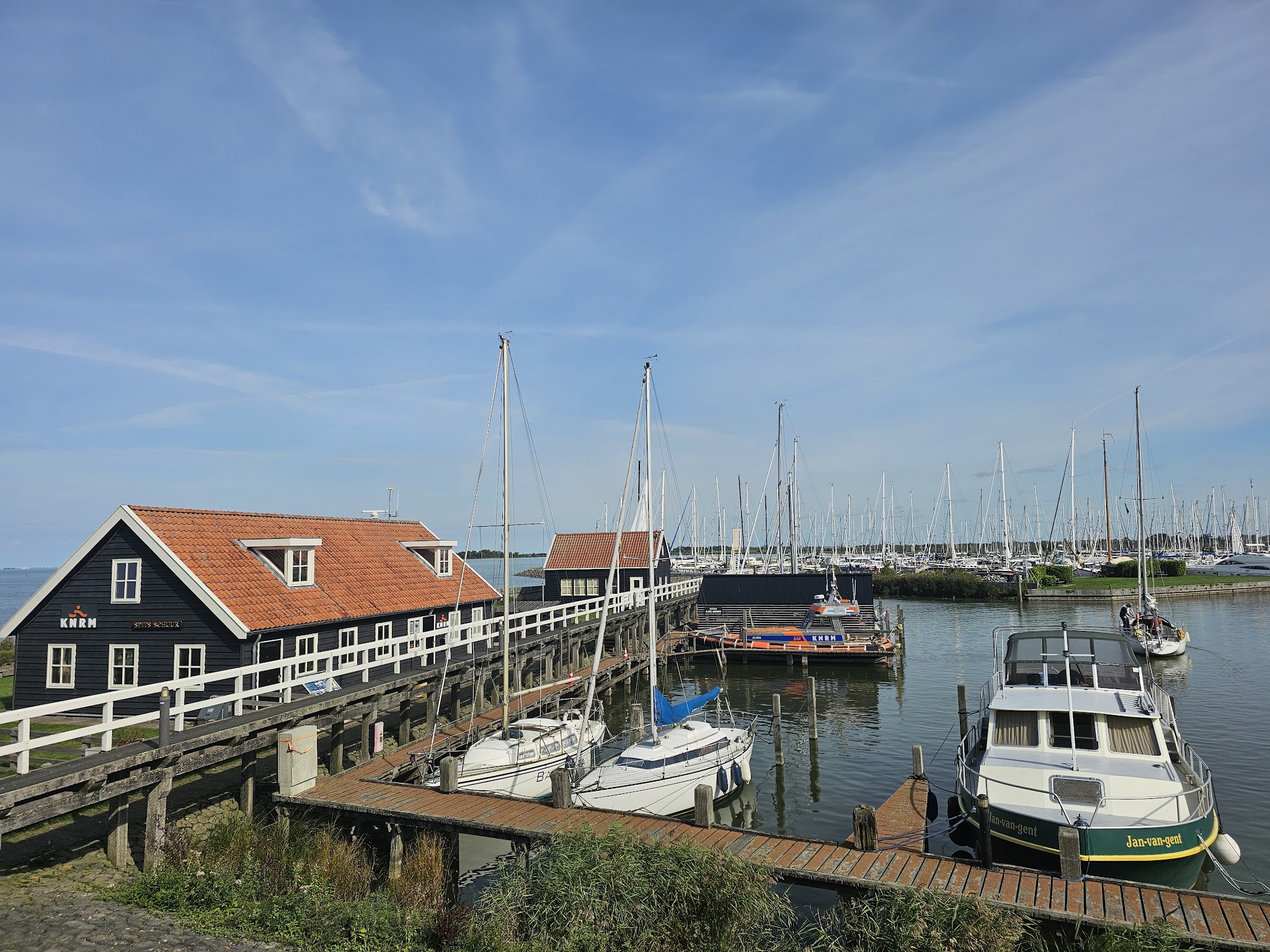Kleiner Hafen mit Holzsteg, rotem Dachhaus und vielen festgemachten Segelbooten unter klarem Himmel.
