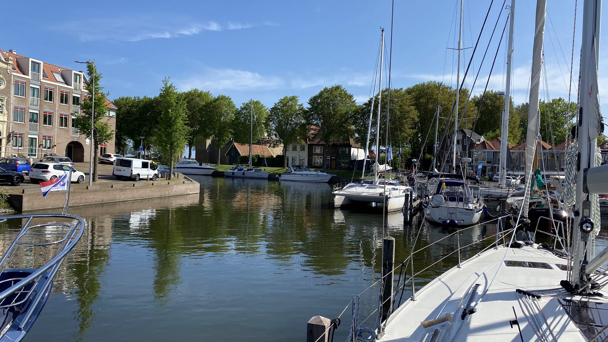 Ruhiger Stadthafen mit angelegten Segelbooten, umgeben von Backsteinhäusern, Autos und grünen Bäumen unter blauem Himmel