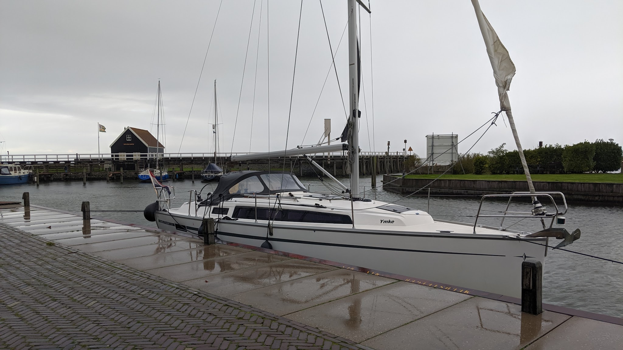 Weißes Segelboot namens „Ymke“ liegt an regennasser Kaimauer in ruhigem Hafen, grauer Himmel