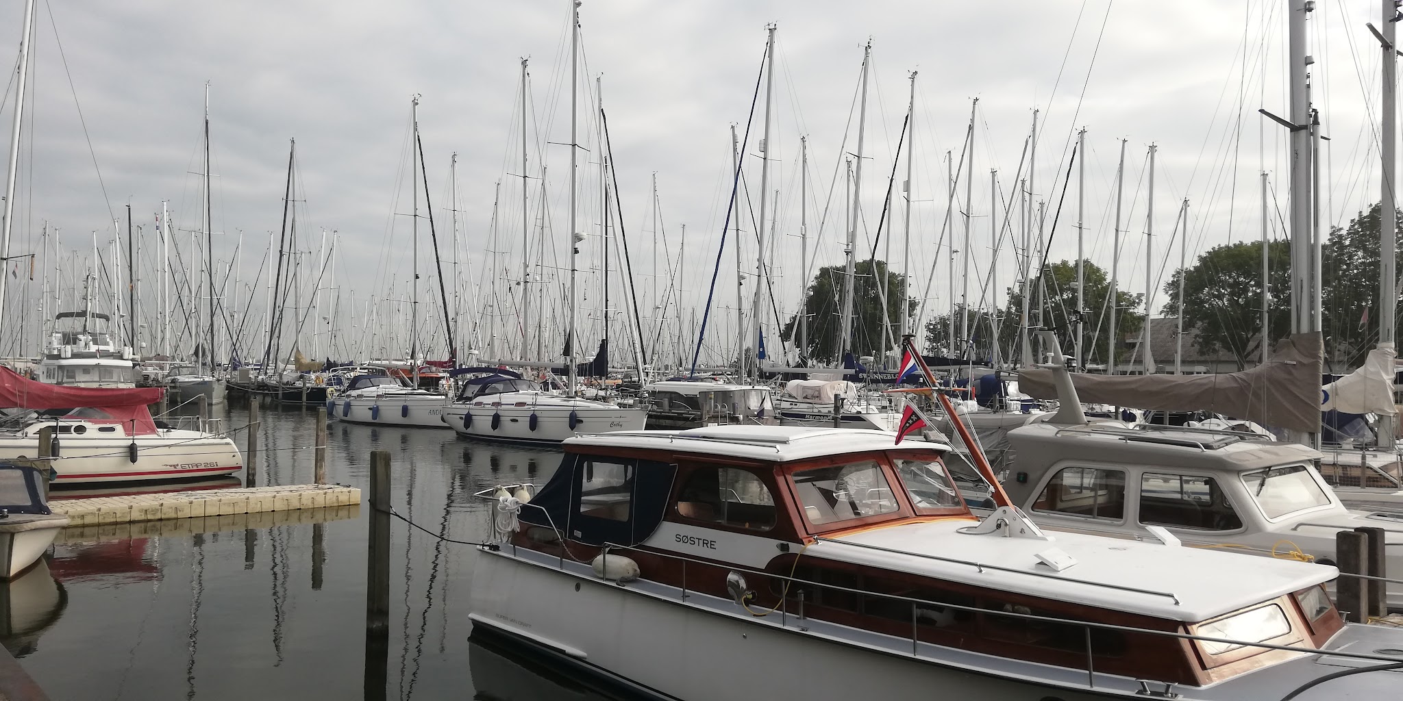Hafen voller Segelboote; im Vordergrund ein klassisches Motorboot mit Flagge, Spiegelungen im ruhigen Wasser.