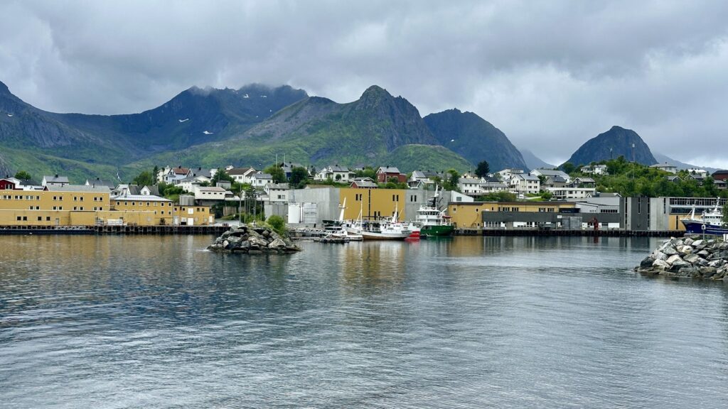 Kleiner nordischer Hafen mit gelben Lagerhäusern, Fischerbooten und weißen Häusern vor grünen, wolkenverhangenen Bergen.