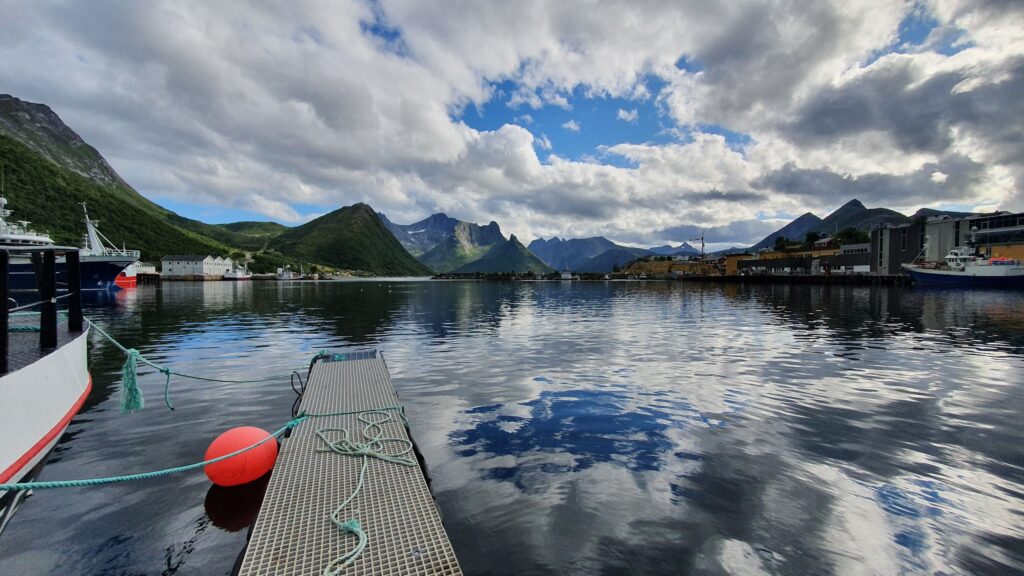Steg mit roter Boje an ruhigem Fjord; Berge, Fischerdorf und teils bewölkter Himmel spiegeln sich im Wasser.