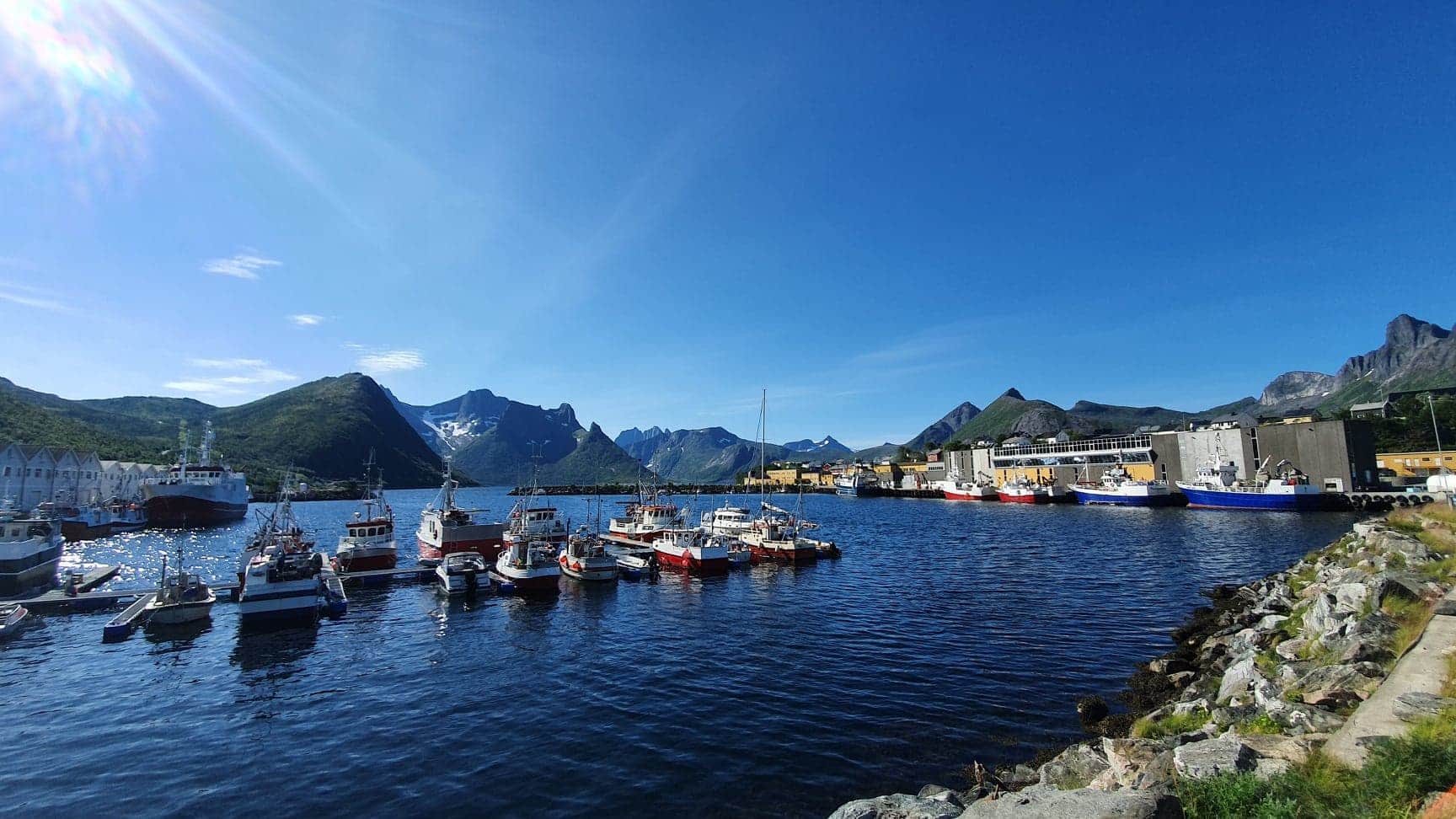 Kleine Fischerboote liegen im klaren Hafen, dahinter grüne Berge mit Gipfeln, alles unter wolkenlosem, strahlend blauem Himmel.