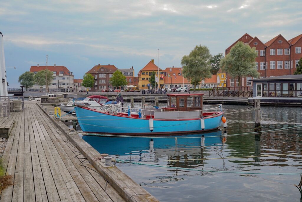 Blaues Fischerboot im ruhigen Hafen vor bunten Backsteinhäusern und Holzsteg
