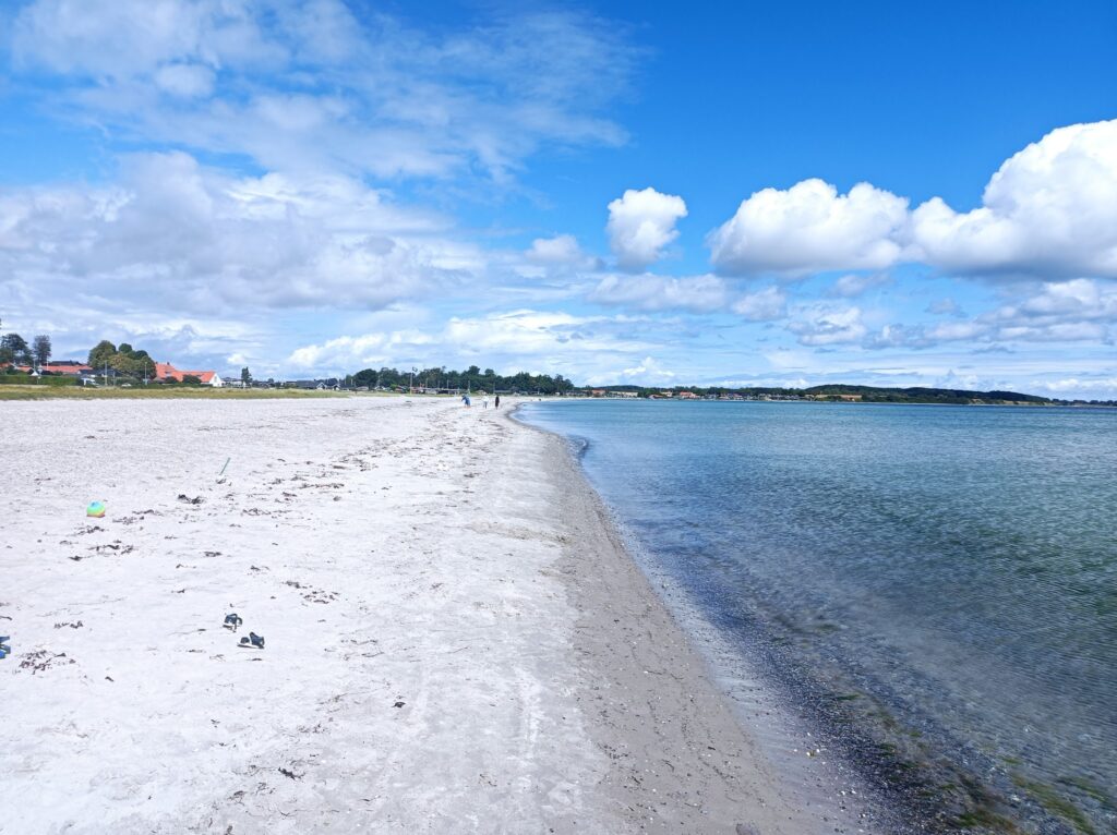 Breiter, heller Strand mit wenigen Spaziergängern, rechts ruhiges türkisblaues Meer, im Hintergrund Dorf mit roten Dächern