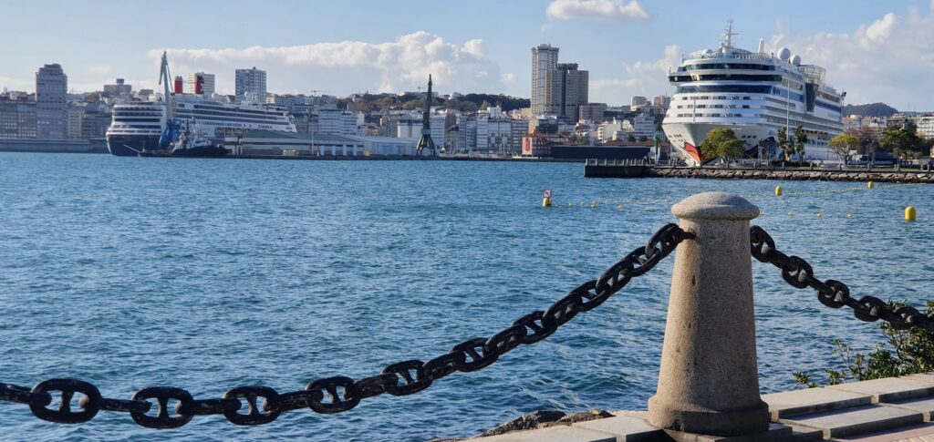 Hafen mit zwei großen Kreuzfahrtschiffen vor Stadtsilhouette, im Vordergrund Poller mit schwerer Kette über blauem Wasser.