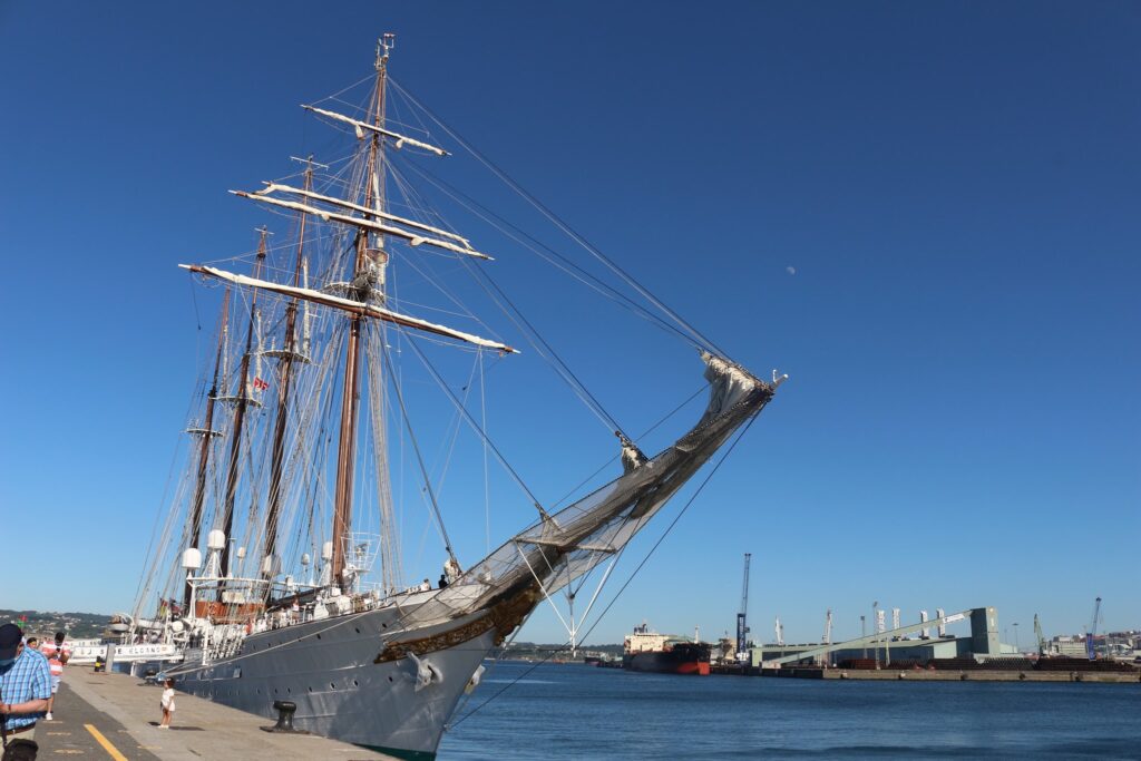 Großes Segelschiff mit hohen Masten im Hafen vertäut; klarer Himmel, wenige Passanten am Kai.