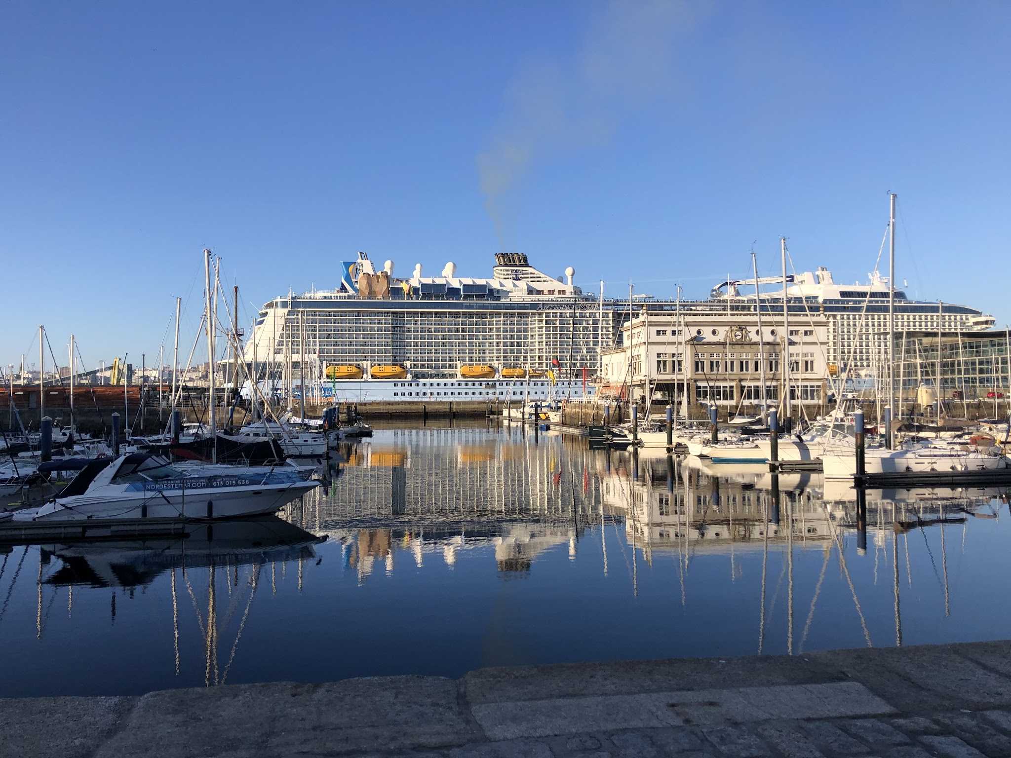 Ruhiger Hafen mit vielen Segelbooten, großes Kreuzfahrtschiff dahinter; klares Wasser spiegelt Schiffe und blauen Himmel