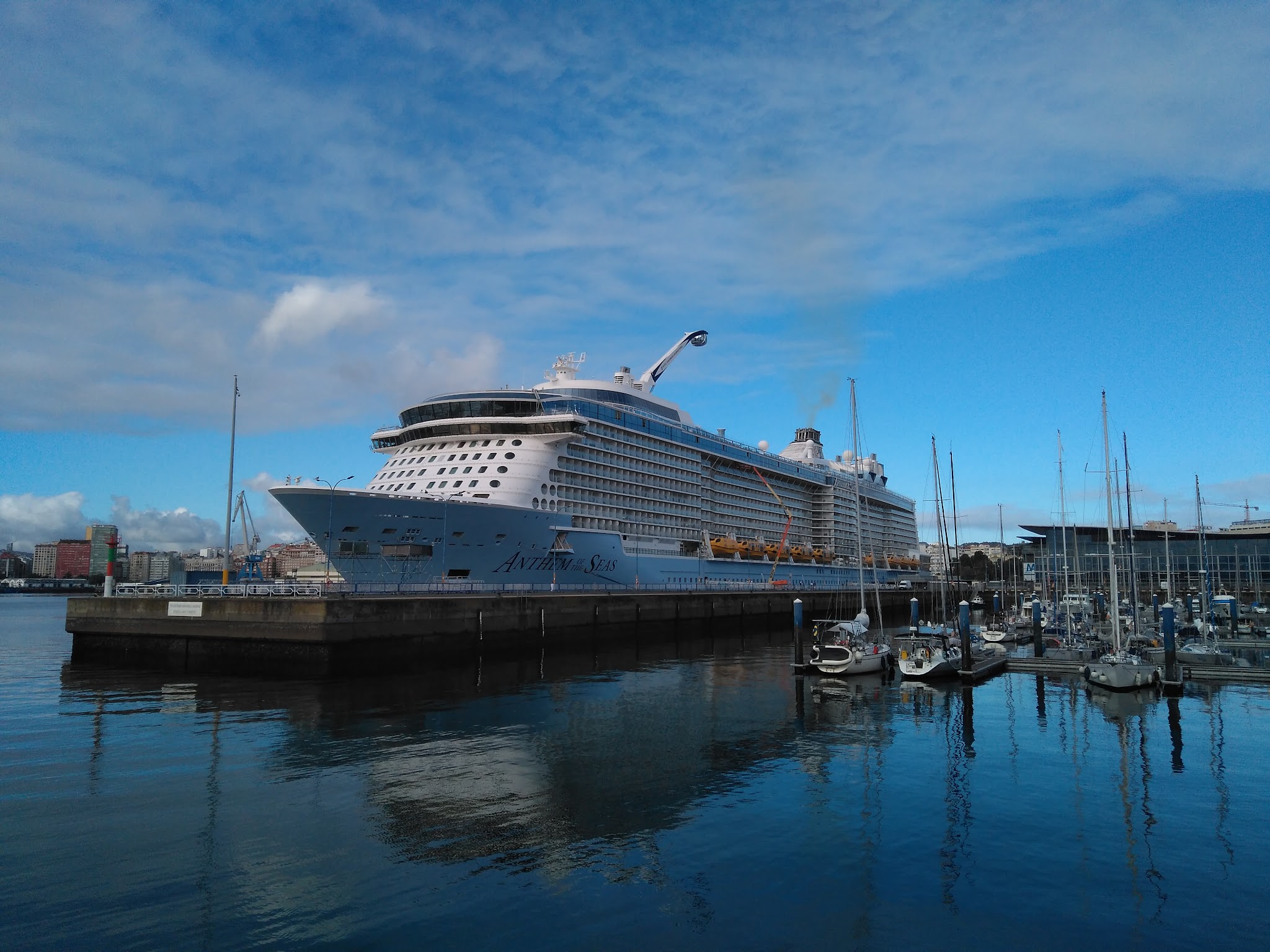 Großes Kreuzfahrtschiff am Pier; daneben kleine Segelboote im Hafen unter blauem Himmel.