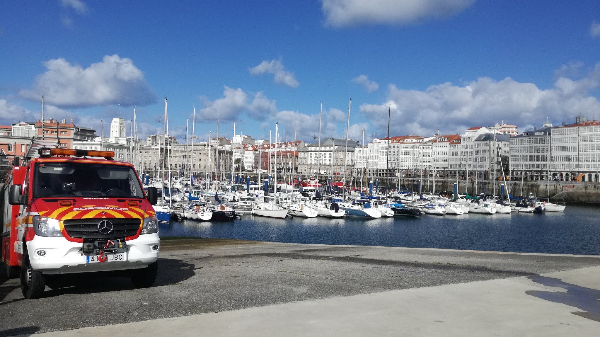 Rotes Einsatzfahrzeug am Kai, dahinter Yachthafen voller Segelboote vor heller Altstadt und blauem Himmel.